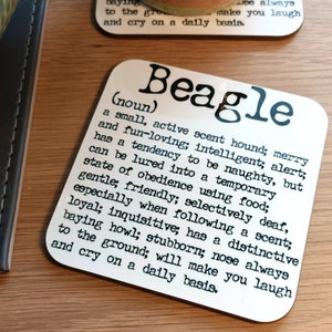 May include: White ceramic coaster with black text defining the word "Beagle". The text describes the Beagle as a small, active scent hound, merry and fun-loving, intelligent, alert, and has a tendency to be naughty, but can be lured into a temporary state of obedience using food.