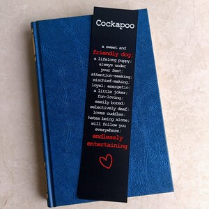 May include: A black bookmark with white text describing the characteristics of a Cockapoo dog. The text includes phrases like "friendly dog", "lifelong puppy", "attention-seeking", "mischief-making", "energetic", "fun-loving", "easily bored", "selectively deaf", "loves cuddles", "hates being alone", "will follow you everywhere", "endlessly entertaining". A red heart is at the bottom of the bookmark.