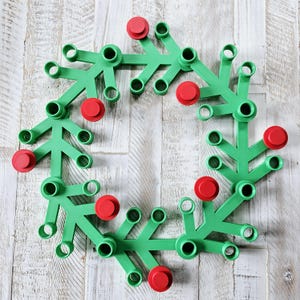 May include: A green plastic wreath with red circular accents. The wreath is composed of interconnected, branch-like segments, each with a circular hole and a red, button-like detail. The design is set against a light wood-grain background.
