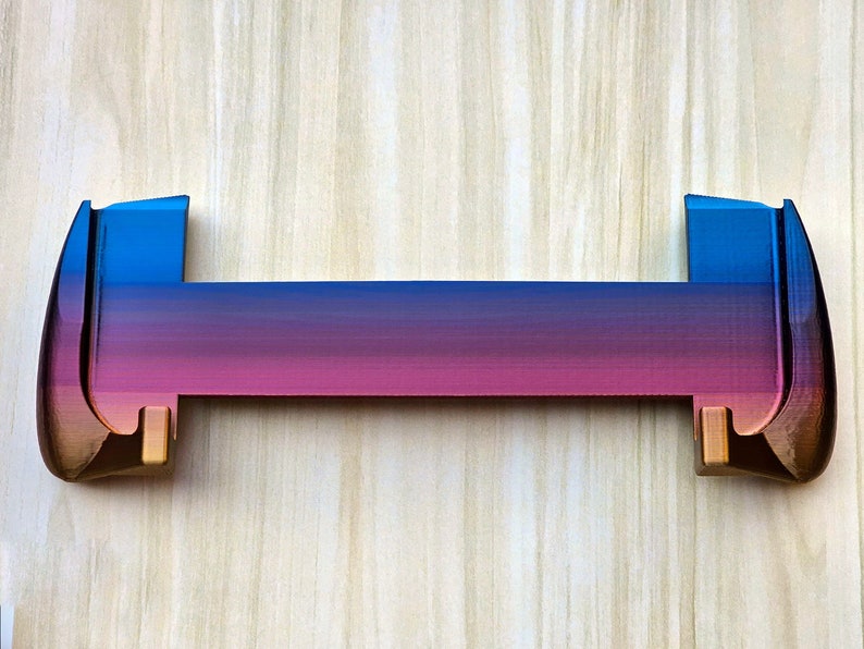 Puede incluir: Un objeto de pl&aacute;stico impreso en 3D, de dos piezas, con un degradado de colores azul, morado y rosa. El objeto tiene una forma rectangular redondeada con una parte superior plana y dos extremos redondeados.