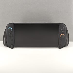 May include: A black handheld gaming console with a large, rectangular screen. The console has attached controllers on either side, with blue and orange accents on the joysticks. The device is resting on a white surface against a gray background.