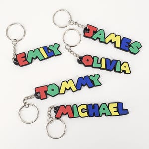 May include: Five colorful keychains with names spelled out in block letters. The names are: Emily, James, Olivia, Tommy, and Michael. Each keychain has a silver key ring.