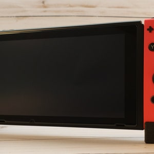 Puede incluir: Una consola Nintendo Switch negra con mandos Joy-Con azules y rojos. La consola est&aacute; en modo port&aacute;til con la pantalla orientada hacia adelante.
