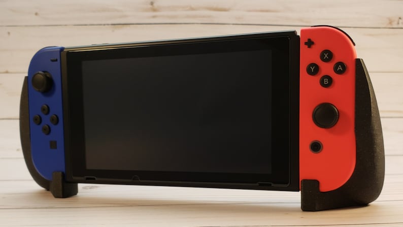 Puede incluir: Una consola Nintendo Switch negra con mandos Joy-Con azules y rojos. La consola est&aacute; en modo port&aacute;til con la pantalla orientada hacia adelante.