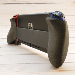Puede incluir: Una consola Nintendo Switch negra con un accesorio de agarre negro y rojo. La consola tiene un bot&oacute;n azul y negro en el lado derecho.