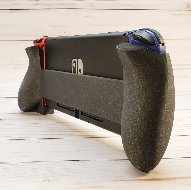 Puede incluir: Una consola Nintendo Switch negra con un accesorio de agarre negro y rojo. La consola tiene un bot&oacute;n azul y negro en el lado derecho.
