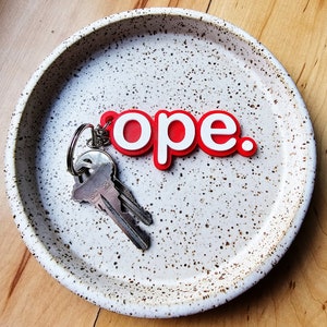 May include: A white ceramic dish with brown speckles, featuring a red and white keychain with the word "ope."  The keychain is hanging on the edge of the dish, and a set of keys is lying on the bottom of the dish.