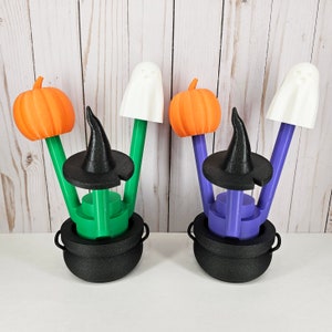 May include: Two Halloween-themed cauldron candle holders, each with a witch's hat, a pumpkin, and a ghost. The cauldron on the left is green and the cauldron on the right is purple.