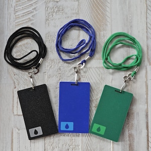 May include: Three ID badge holders with lanyards. One black, one blue, and one green badge. Each badge has a matching lanyard and a silver-colored clasp. Each badge has a white square with a blue water droplet icon.