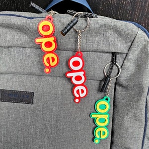 May include: Three keychains with the word "ope" written on them in different colors. The keychains are attached to a gray backpack.