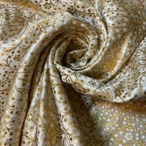 Op de afbeelding: Witte stof met een gouden bloemenpatroon. De stof is zacht en luxueus, perfect voor bekleding of gordijnen.