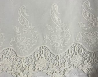 May include: A white fabric featuring an intricate embroidered pattern of repeating paisley motifs and floral elements. The lower edge is finished with a scalloped, openwork lace border that displays a detailed, interconnected floral design.