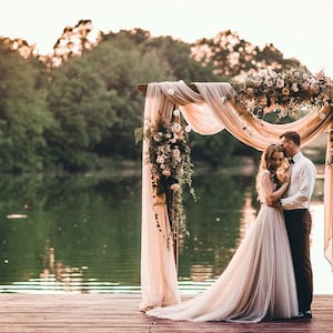 May include: A wedding ceremony setting with a wooden arch decorated with flowers and draped fabric. A couple stands embracing on a wooden dock overlooking a lake. The scene is bathed in warm, natural light, creating a romantic atmosphere.