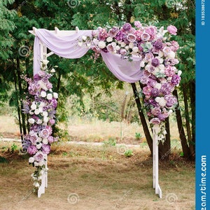 May include: A white wooden wedding arch decorated with an abundance of purple and white flowers and draped with a sheer lavender fabric. The arch is set outdoors in a natural setting, creating a romantic atmosphere.