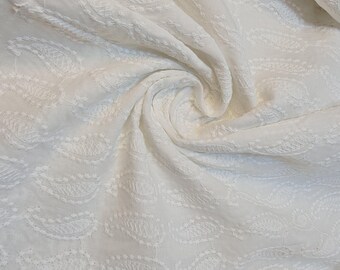 May include: A white fabric featuring an all-over embroidered paisley pattern. The material is gathered in the center, creating soft folds and ripples that highlight the texture of the white-on-white stitching throughout the textile surface.