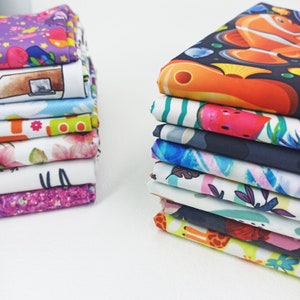May include: A stack of colorful fabric squares with various patterns, including a purple fabric with stars, a blue fabric with a house, a white fabric with flowers, and a black fabric with a fish.