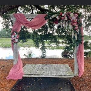 May include: A wooden wedding arch with a platform, draped with pink fabric and adorned with floral arrangements. The arch is set outdoors with a lake and trees in the background, creating a natural and romantic setting.