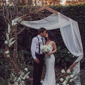 May include: A couple kissing under a wooden wedding arch draped with white fabric. The bride wears a white lace gown and holds a bouquet. The arch is decorated with flowers and greenery, set in a garden setting.