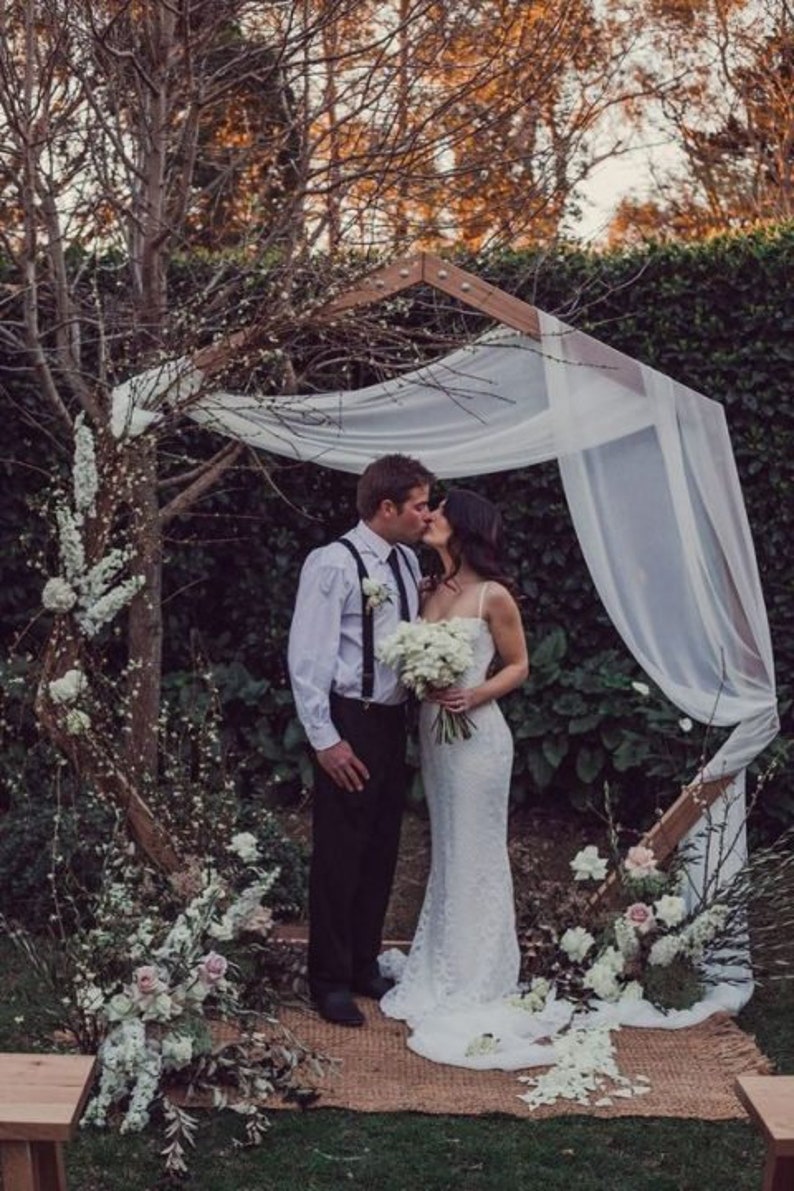 May include: A couple kissing under a wooden wedding arch draped with white fabric. The bride wears a white lace gown and holds a bouquet. The arch is decorated with flowers and greenery, set in a garden setting.
