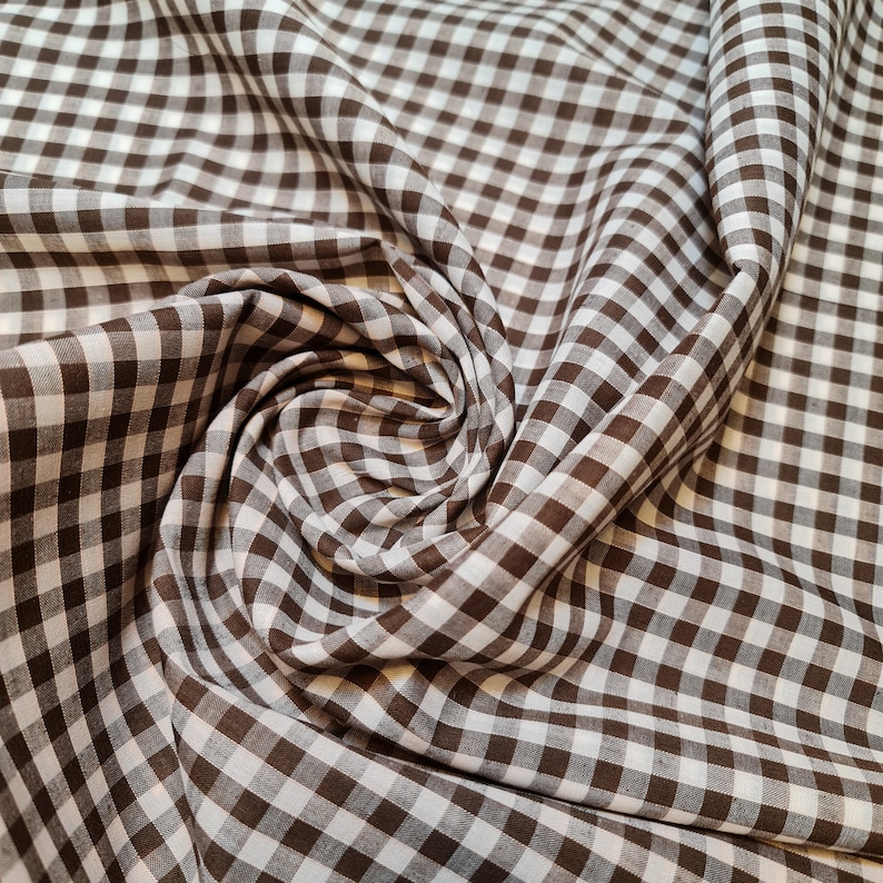 May include: Brown and white gingham fabric with a checkered pattern. The fabric is folded and draped, creating a soft, textured look.