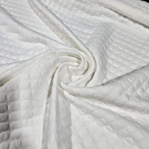May include: A bright white fabric featuring a repeating embossed circular pattern across its surface. The material is draped in soft, flowing folds, highlighting the textured, raised geometric design and the consistent matte finish of the textile.