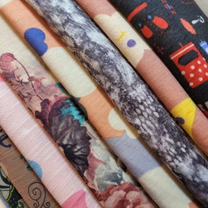 Può includere: Una collezione di campioni di tessuto in vari colori e motivi, tra cui disegni floreali, geometrici e astratti. I campioni sono disposti in una pila, con il campione superiore che è un tessuto nero con pois bianchi.