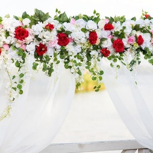 May include: A white wedding arch adorned with vibrant red and white flowers, lush greenery, and cascading white floral accents. The arch is draped with flowing white fabric, creating an elegant and romantic setting for a ceremony.