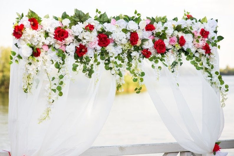 May include: A white wedding arch adorned with vibrant red and white flowers, lush greenery, and cascading white floral accents. The arch is draped with flowing white fabric, creating an elegant and romantic setting for a ceremony.