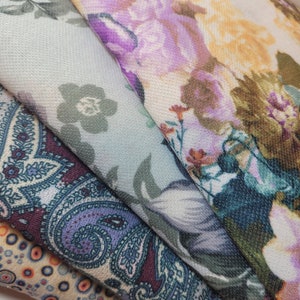 Pode incluir: Quatro amostras de tecido com padrões florais e paisley. As amostras são sobrepostas, com a amostra superior sendo um tecido azul claro com um padrão floral. A segunda amostra é um tecido floral roxo e branco. A terceira amostra é um tecido paisley marrom e azul. A amostra inferior é um tecido bege com um padrão geométrico.