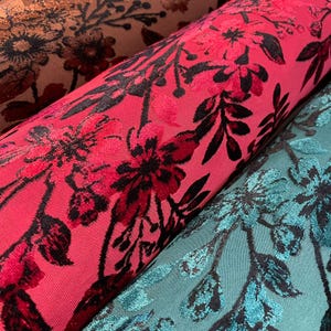 May include: Close-up of rolled fabric with floral patterns. The fabric features a vibrant red background with black and dark red floral designs. Another roll of fabric in teal with black floral patterns is visible, along with a brown fabric with dark floral patterns.