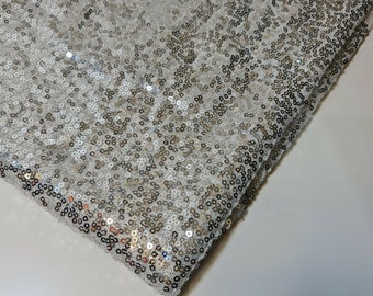 May include: A close-up of a white fabric with silver sequins. The fabric is folded in half, showing the sequins on both sides.