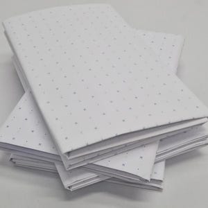 May include: A stack of folded white fabric sheets featuring a repeating pattern of small, dark blue cross symbols. The fabric is neatly layered, showing the crisp white material and the consistent geometric print across the surface of each piece.