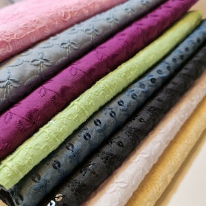 May include: A stack of eight fabric swatches in various colours and textures, including pink, light blue, purple, green, dark blue, black, white, and beige. Each swatch features a delicate embroidered floral pattern.