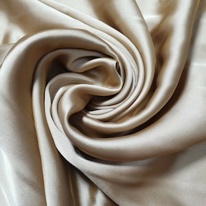 May include: A close-up of a smooth, silky fabric in a light beige color. The fabric is draped and folded, creating soft, flowing curves.
