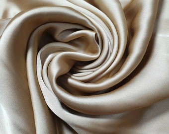 May include: A close-up of a smooth, silky fabric in a light beige color. The fabric is draped and folded, creating soft, flowing curves.