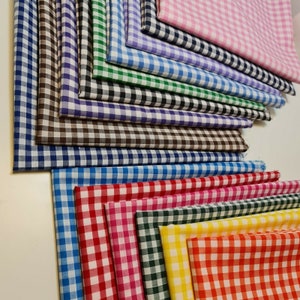 Op de afbeelding: Een stapel kleurrijke gingham-stoffen in verschillende kleuren, waaronder roze, blauw, groen, rood, geel en zwart. De stof is gevouwen en gestapeld in een nette stapel.