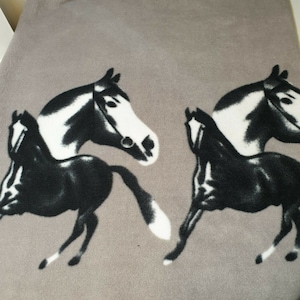 Puede incluir: Una manta de forro polar gris con un patrón repetido de caballos corriendo. Los caballos están representados en blanco y negro, con crines y colas detalladas. La manta es suave y acogedora.