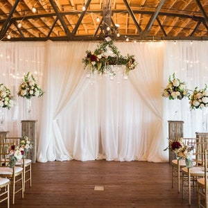 May include: An indoor wedding ceremony setup with white draping, floral arrangements, and gold chairs. The backdrop features sheer white curtains, with floral arrangements on pedestals and hanging from a chandelier. String lights and a wooden floor complete the scene.