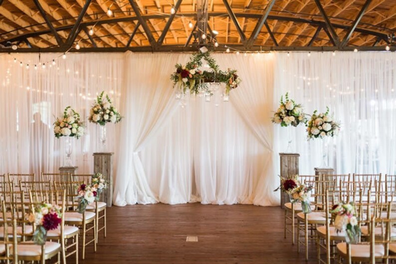 May include: An indoor wedding ceremony setup with white draping, floral arrangements, and gold chairs. The backdrop features sheer white curtains, with floral arrangements on pedestals and hanging from a chandelier. String lights and a wooden floor complete the scene.