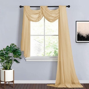 May include: Beige sheer window curtains with a swag valance. The curtains are draped over a black rod, framing a window with a view of trees. A potted plant sits to the left, and a framed abstract art piece hangs on the wall.