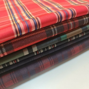 May include: A stack of plaid fabric in various colors, including red, black, and green. The fabric is folded neatly and has a soft, textured feel.