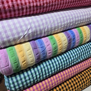 Puede incluir: Una pila de rollos de tela con varios patrones y colores. El rollo superior es de cuadros vichy rojos y blancos, seguido de uno lavanda y blanco, una tela de rayas multicolor, uno de cuadros verde azulado y blanco, y uno amarillo y negro.