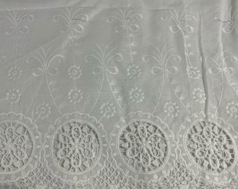 May include: A white fabric featuring intricate embroidery with floral motifs and scrolling vines. The bottom edge is finished with a wide, circular lace border pattern and a delicate scalloped trim with small, rounded pom-pom accents.