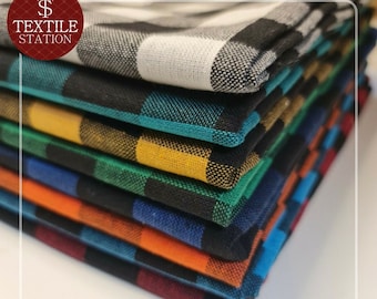 May include: A stack of eight plaid fabric swatches in various colors, including black, white, teal, yellow, green, blue, orange, and red. The fabric is folded neatly and the top swatch is white and black.