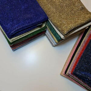 Puede incluir: Una pila de tela brillante en varios colores, incluyendo dorado, verde, azul y rojo. La tela está dispuesta en una pila, con la capa superior dorada.