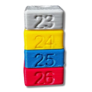 Könnte beinhalten: Ein Stapel aus vier bunten Blöcken, jeder mit einer anderen Nummer bedruckt. Die Blöcke sind grau, gelb, blau und rot, und die Zahlen sind 23, 24, 25 und 26.