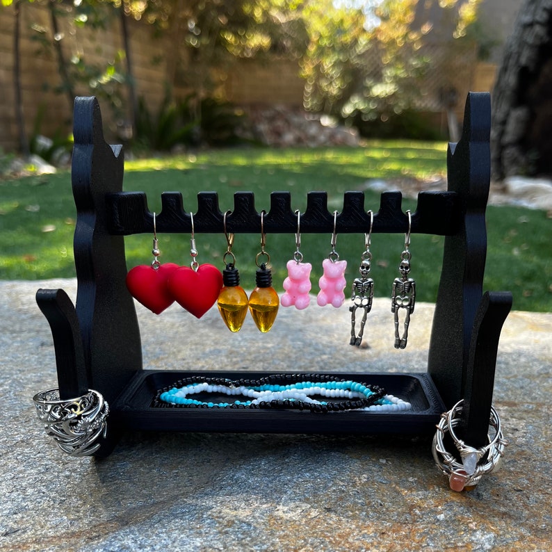 May include: A black jewelry stand with six pairs of earrings. The earrings are a variety of shapes and colors, including hearts, light bulbs, and gummy bears. There is also a black and white beaded necklace on the stand.