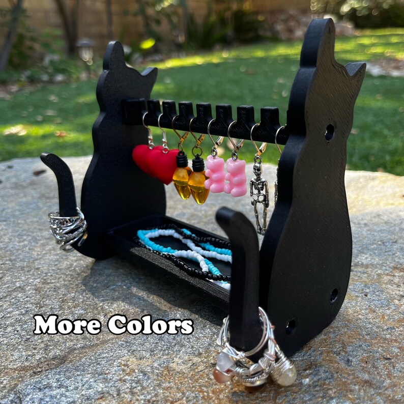 May include: Black cat-shaped jewelry organizer with a shelf for rings and a hanging rack for earrings. The rack has a heart, light bulbs, gummy bears, and a skeleton charm hanging from it. The text "More Colors" is visible in the image.