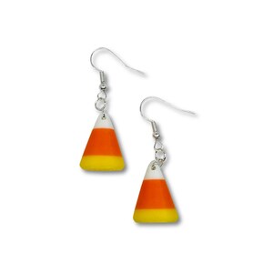 May include: A pair of dangling earrings featuring a candy corn design. Each earring has a silver-colored fish hook and a charm shaped like a candy corn. The candy corn charms are white, orange, and yellow.