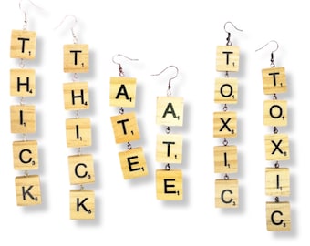 May include: Three pairs of dangling earrings made from light-colored wooden letter tiles connected by silver-toned metal chains. The tiles spell out the words THICK, HATE, and TOXIC, with each letter tile featuring a black letter and a small numerical value.